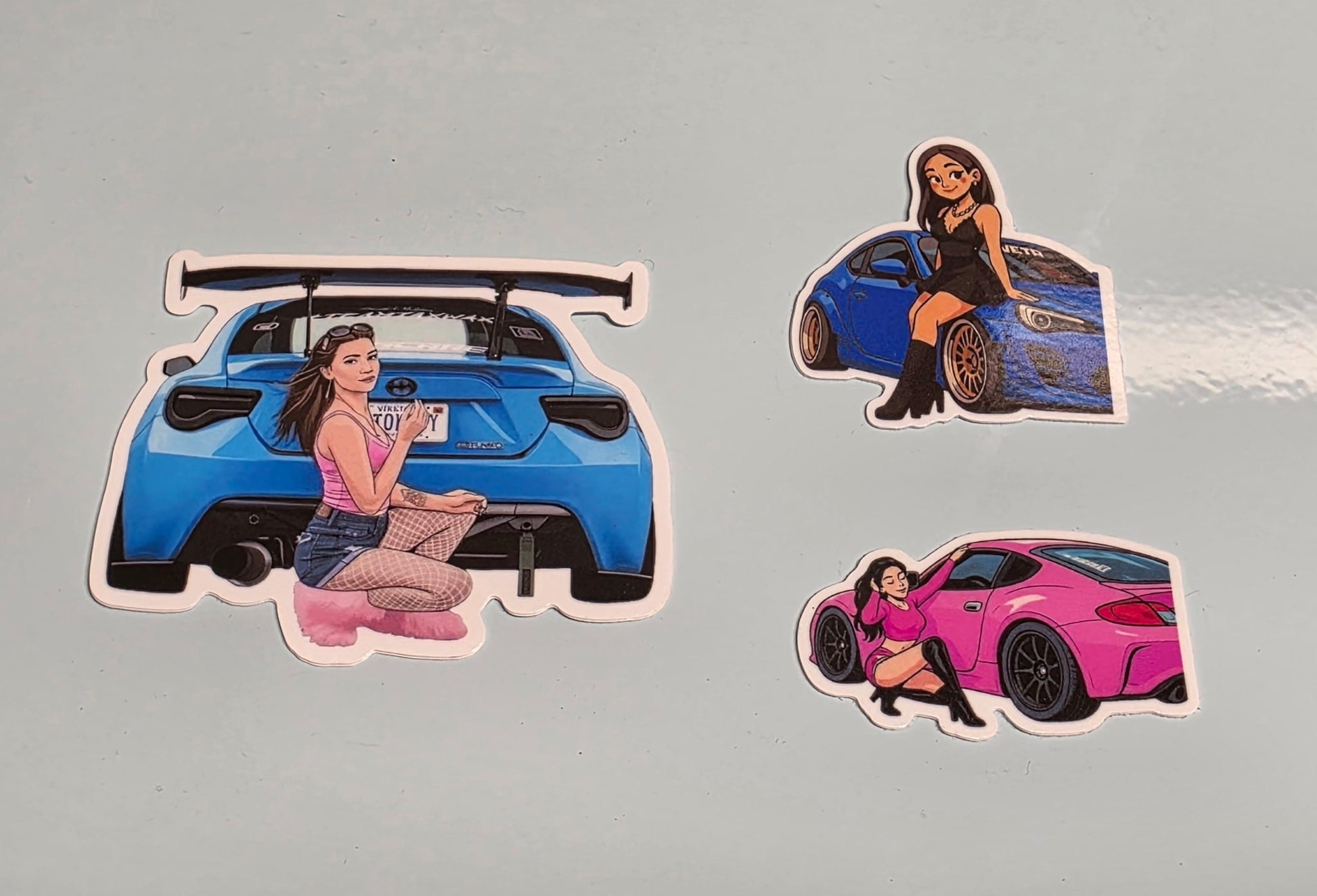 Stickers of a woman with a blue car, a woman with a pink car, and a woman with a black car on a gray background.
